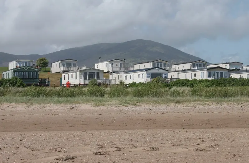 White static caravans behind a sandy beach, with a large hill rising behind them